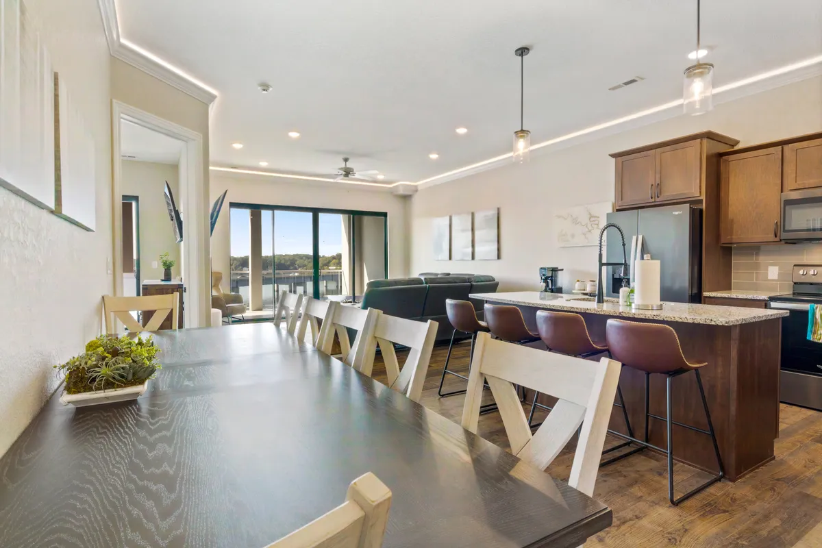 This image captures a spacious open-plan living area that seamlessly flows into a dining space and kitchen. The room is filled with natural light pouring in through large windows, offering beautiful lake views. The living area is arranged for relaxation and social gatherings, with a cozy dining table ready for shared meals.