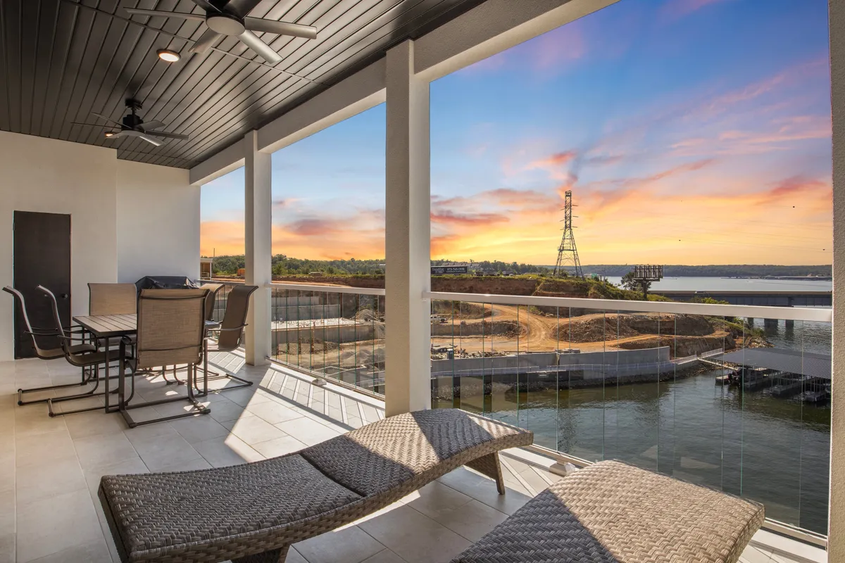 A beautifully appointed private balcony featuring comfortable seating, a dining table set for alfresco meals, and an expansive water view framed by a glowing sunset sky. The atmosphere is tranquil and elegant, perfect for outdoor dining or quiet reflection while watching boats drift by.