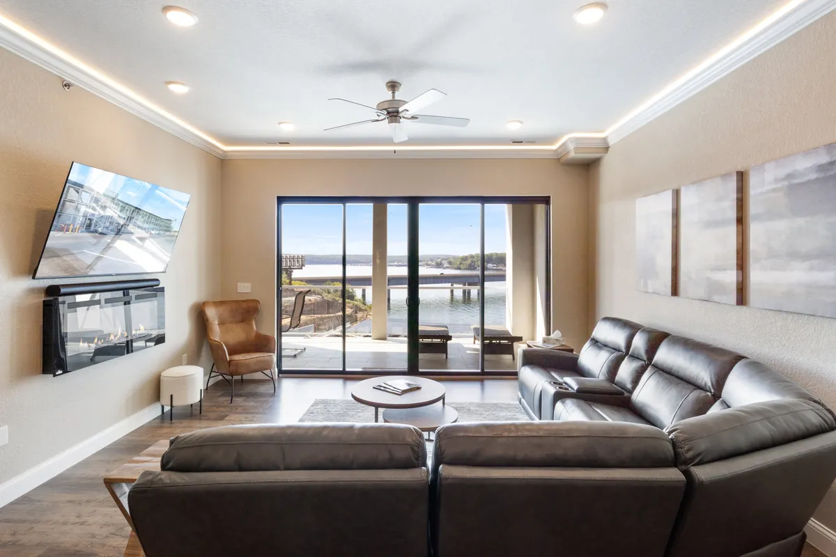 The focus is on a modern leather sectional sofa facing a flat-screen TV, with expansive glass doors providing uninterrupted views of the lake beyond. Stylish furnishings and artwork create a contemporary, welcoming atmosphere ideal for movie nights and downtime with friends or family.