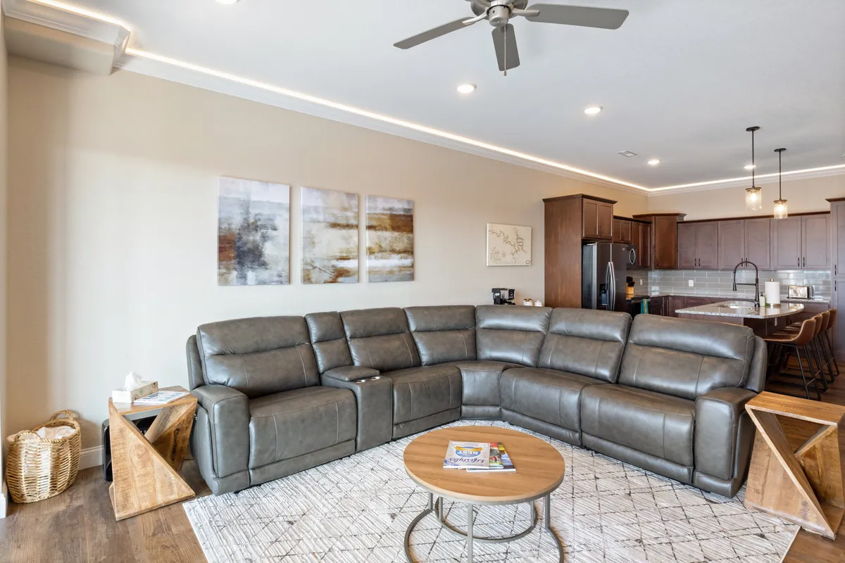 A cozy, open-concept living room with a large leather sectional, round coffee table, wall art, and a kitchen area in the background. The inviting space is staged for family movie nights or board games after a day of local adventures.