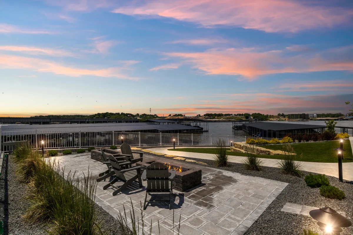 A cozy fire pit surrounded by Adirondack chairs faces a stunning water view, with the sky painted in sunset hues. The setup invites groups to gather, socialize, and enjoy the warmth of the fire against a beautiful natural backdrop.