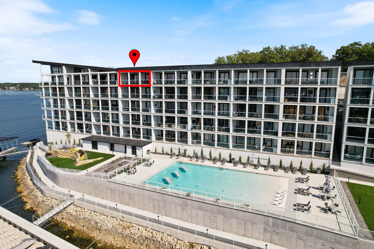 The image shows a sparkling lakeside swimming pool in the foreground, perfect for both relaxing swims and social gatherings, with the condo's modern facade and expansive windows rising behind it. Beyond the pool, stunning panoramic views of the lake stretch out to the horizon, with boats passing by and lush greenery lining the water's edge. The scene is bathed in soft, golden-hour light, evoking a tranquil and inviting mood.