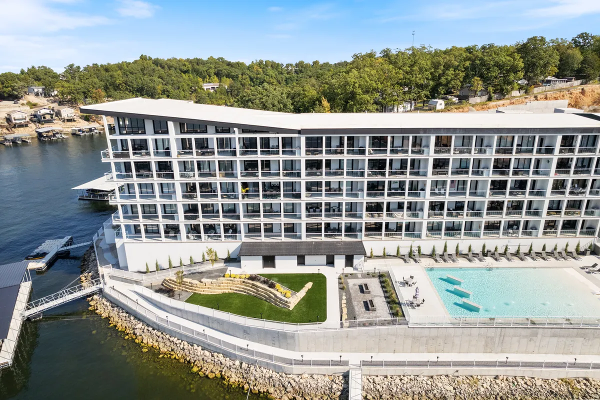 A wide exterior shot featuring a modern hotel building set right on the water, with a sparkling swimming pool, landscaped green areas, lounge chairs, and visible boat docks. The scene is active yet tranquil, capturing both relaxation and direct access to boating amenities.