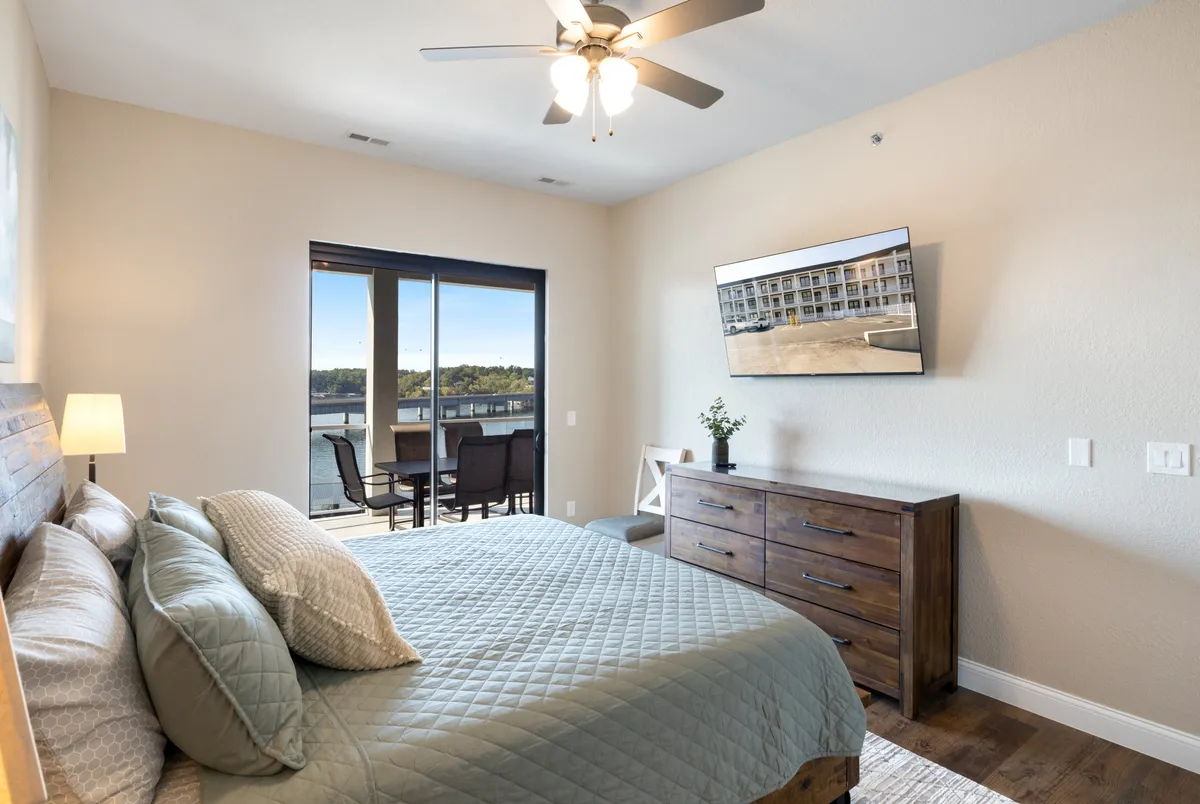 A cozy and elegantly decorated king bedroom with a quilted bedspread, large window offering scenic views, a wooden dresser with a small plant, and a wall-mounted TV. The soft lighting and careful accents make the room inviting and restful.