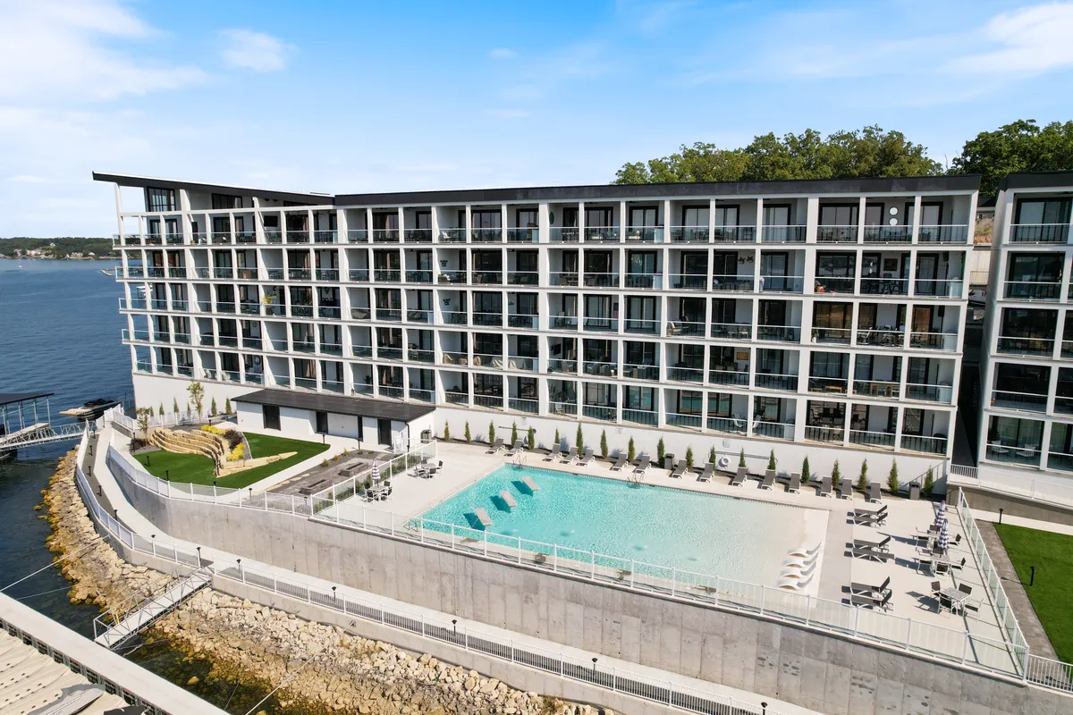 This image displays a sparkling lakeside swimming pool adjacent to a modern hotel building, with a dock extending into the lake and views of the surrounding landscape. The scene captures people lounging by the water, inviting a sense of relaxation and enjoyment for all ages.