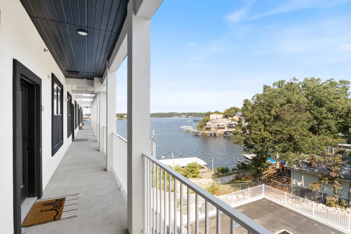 A tranquil balcony overlooks the lake, bordered by stylish railings and offering sweeping water views. Neighboring houses are visible in the distance, and the space is set up for outdoor relaxation, socializing, or enjoying meals al fresco.